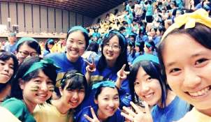 ホールで青や黄色のハチマキをした大勢の学生たちがおり、そのうちの9名の女性が手前のカメラに向かって集合している